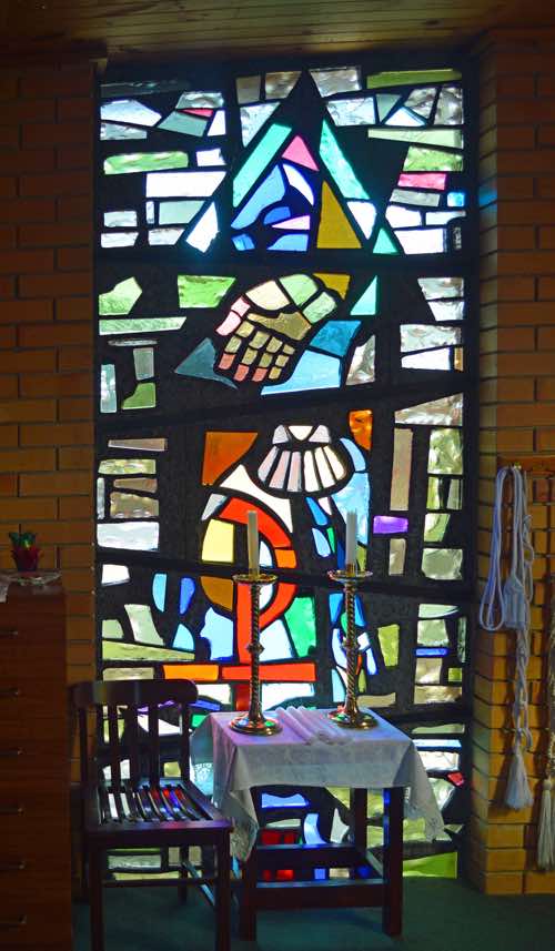 The small square chapel in the south-eastern corner has two stained glass windows of modern design.  This window has a God theme: the Chi-Rho symbol for Christ, the Father’s hand reaching down, the tail (at least) of the Dove of the Holy Spirit.  An upward arrow suggests man’s seeking after God.