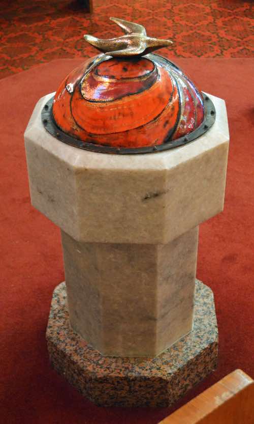 The modern style and orange colouring of the baptismal font and the ambo suggest the work of the same artist.  Atop the domed font cover is a golden dove.