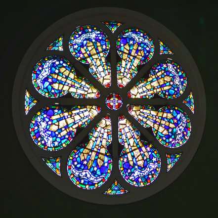 The rose window sits high in the Eastern gable.  With the Cross at the centre, it depicts a sunburst of light reaching out to the stars.  [22]  [Click to ZOOM.]